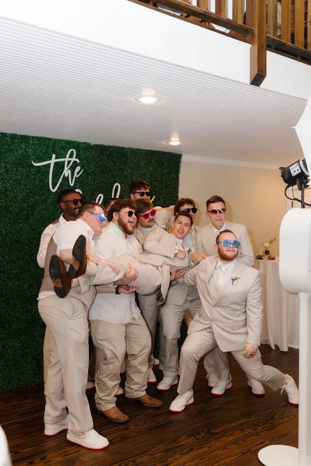 Groomsmen in tan suits hoisting one of their own, all in novelty sunglasses, in front of a green hedge wall.