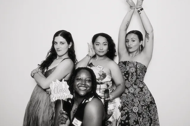 Four women in a black and white booth portrait, one holding a Class Reunion prop sign.