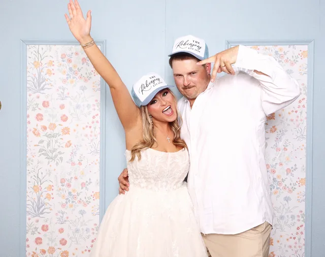 Bride and groom cheering in matching trucker hats, in front of a pale blue wall papered with small flowers.