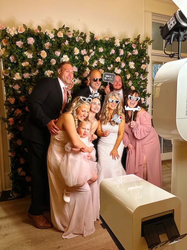 Wedding guests posing with props in front of a pink and peach flower wall, the open air booth beside them.