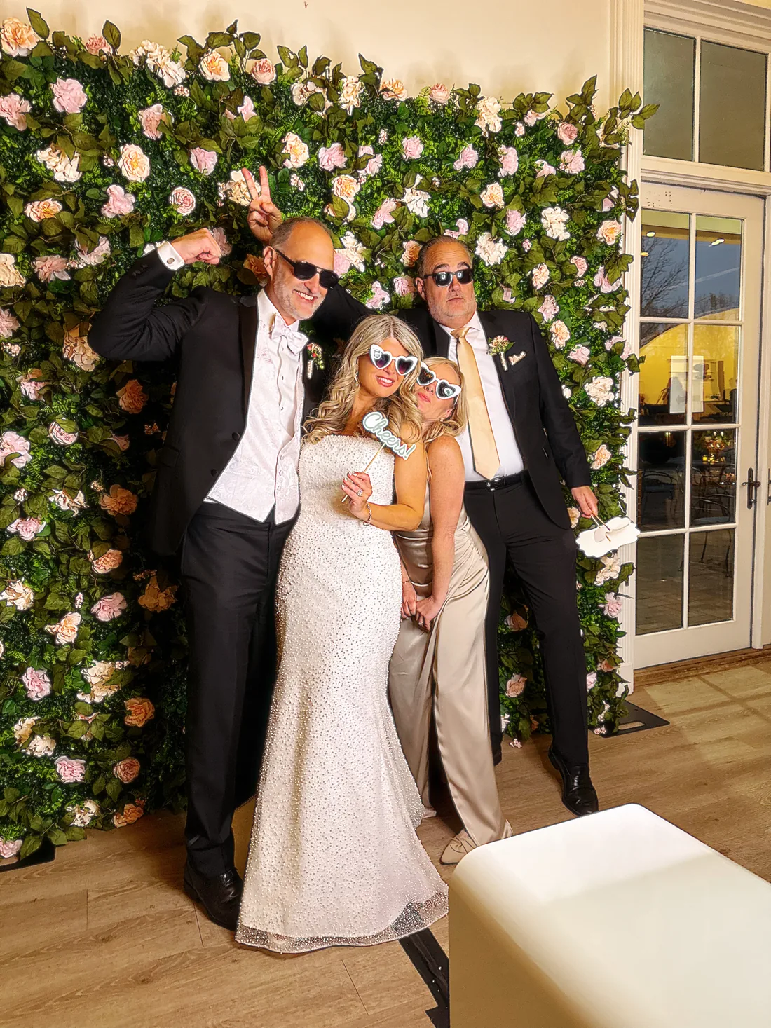 Bride and wedding guests in sunglasses posing in front of the white flower wall backdrop.