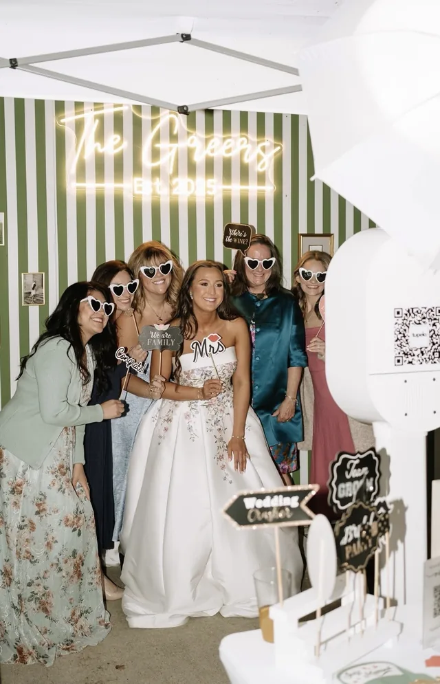 Bride and her guests in heart sunglasses, holding Mrs and We Are Family props, next to the open air booth.