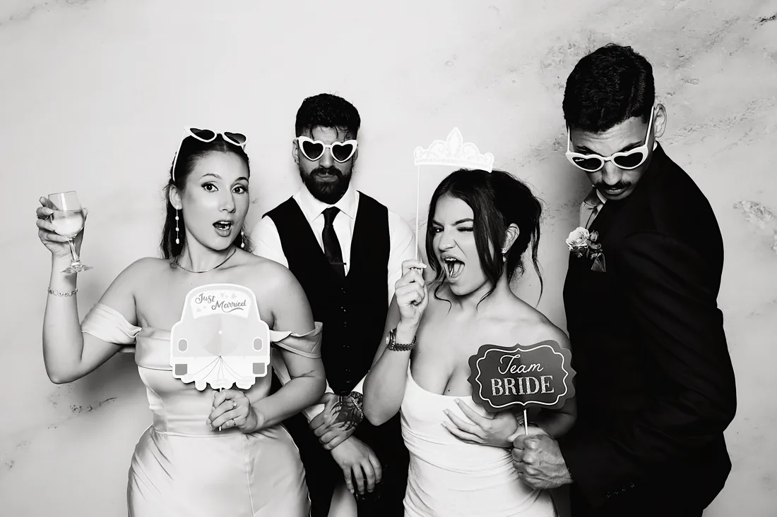 Two couples posing with props in a black and white booth photo.